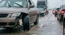 Damaged gold car with detached bumper on a wet highway amidst traffic.