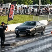 Kyle Loftis, founder of 1320Video, smiles while wearing a black t-shirt with the '1320Video' logo, at an outdoor drag racing event.